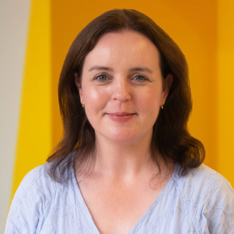 A woman with shoulder-length brown hair smiles softly, wearing a light blue top. She stands in front of a bright yellow background.