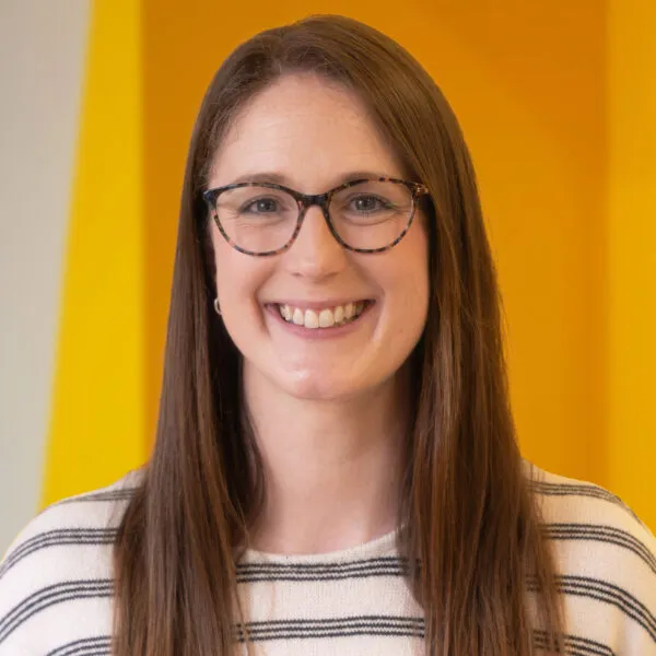 Woman with long brown hair and glasses smiling, wearing a white striped shirt, standing in front of a bright yellow background.