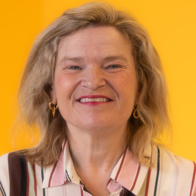 Smiling woman with blonde hair, wearing gold hoop earrings and a striped button-up shirt, standing in front of a solid yellow background.