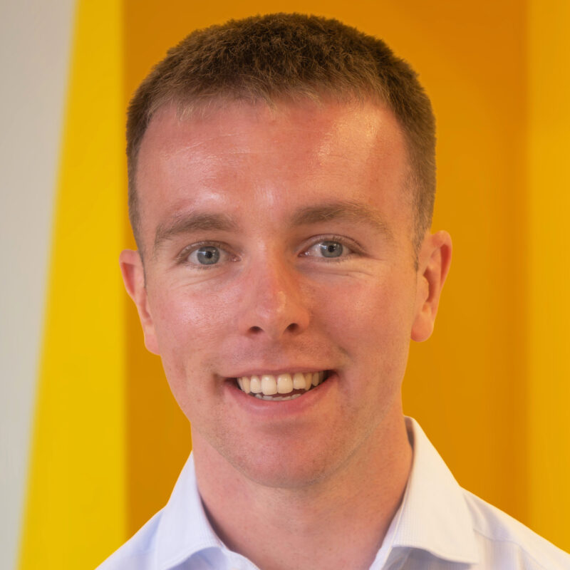 A smiling young man with short brown hair, wearing a light-coloured collared shirt, stands in front of a bright yellow background.