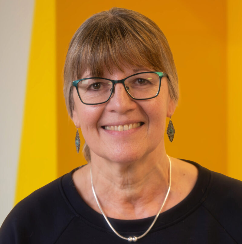 A smiling older woman with glasses, short brown hair, and earrings is standing in front of a bright yellow background. She is wearing a dark top and a silver necklace.