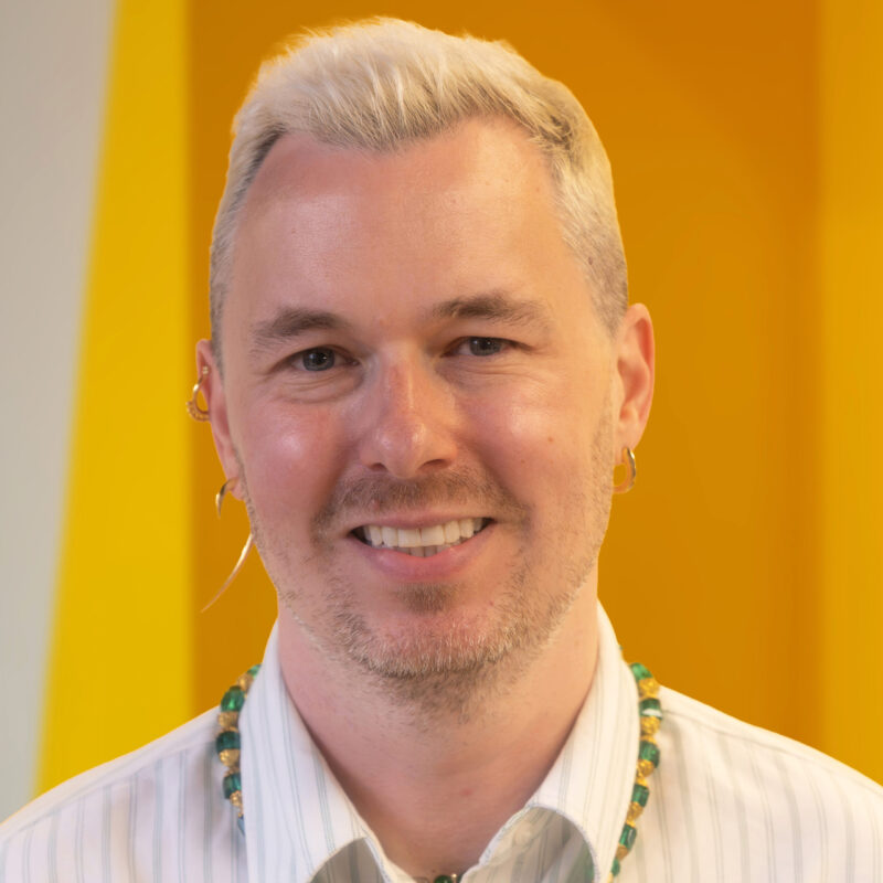 A smiling person with short blond hair, wearing a striped shirt, green beaded necklace, and gold earrings, stands in front of a bright yellow background.