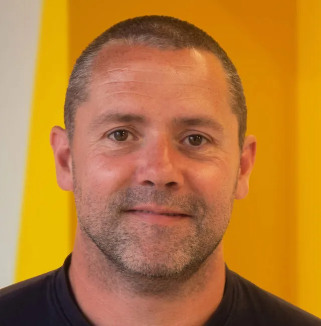 A man with short brown hair and stubble smiles slightly, standing in front of a bright yellow and white background.