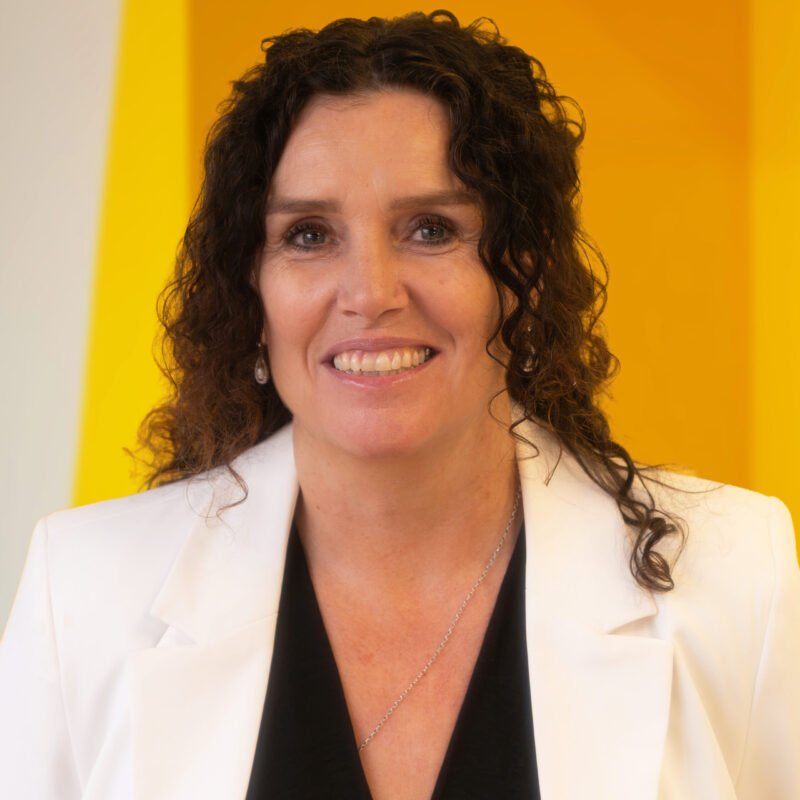 A woman with curly brown hair, wearing a white blazer over a black top, smiles in front of a vibrant yellow background.