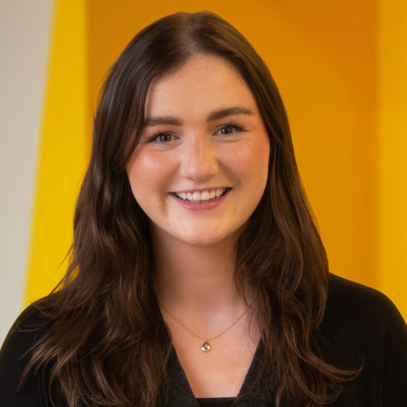 A woman with long brown hair, wearing a black top and a necklace, smiles in front of a yellow background.