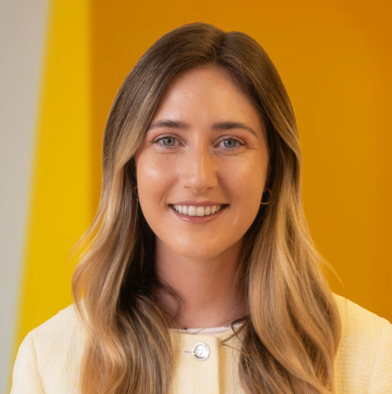 A woman with long blonde hair and a light yellow blazer smiles in front of a vibrant yellow background.