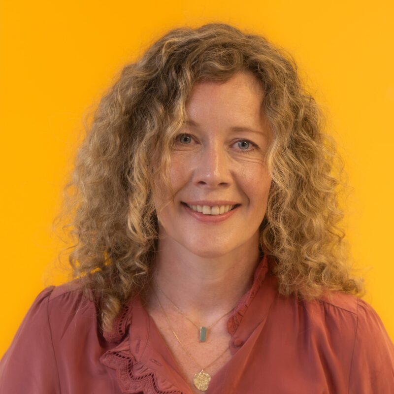 A woman with curly blonde hair wearing a mauve blouse and two necklaces smiles in front of a bright yellow background.