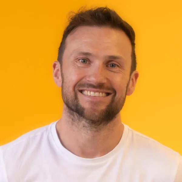 A man with short brown hair and a beard smiles at the camera, wearing a white T-shirt against a bright yellow background.