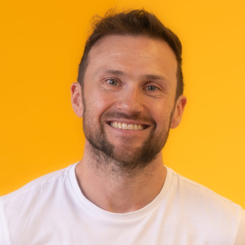 A man with short brown hair and a beard smiles at the camera, wearing a white T-shirt against a bright yellow background.