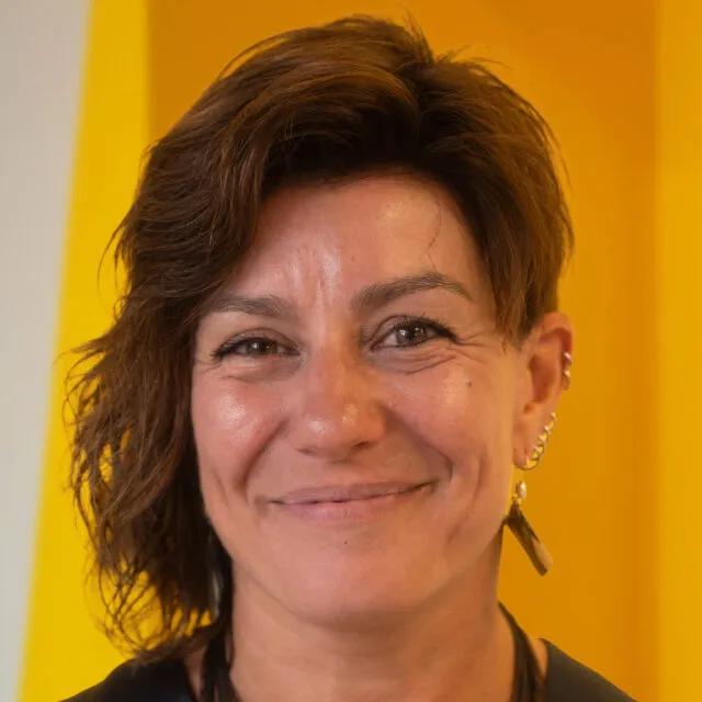 Smiling person with short brown hair, wearing earrings and a black shirt, standing in front of a bright yellow and white background.