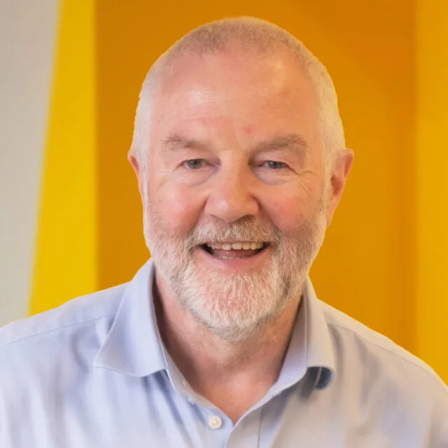 Smiling older man with a white beard wearing a light blue shirt, standing in front of a bright yellow background.