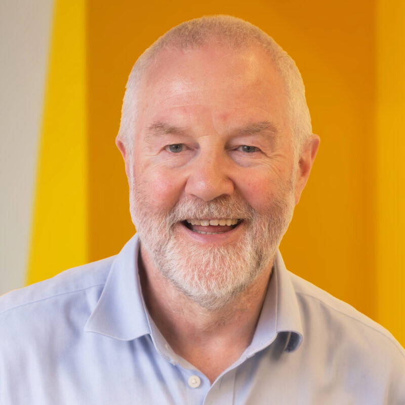 Smiling older man with a white beard wearing a light blue shirt, standing in front of a bright yellow background.