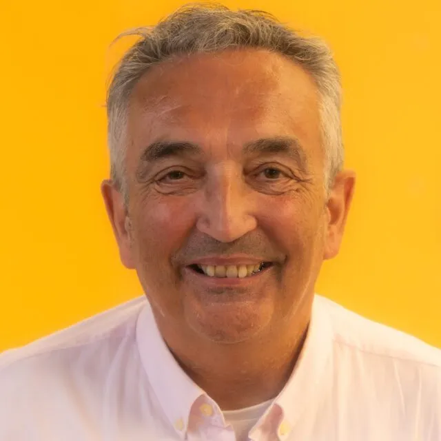 Smiling older man with grey hair wearing a white collared shirt, posed against a bright orange background.