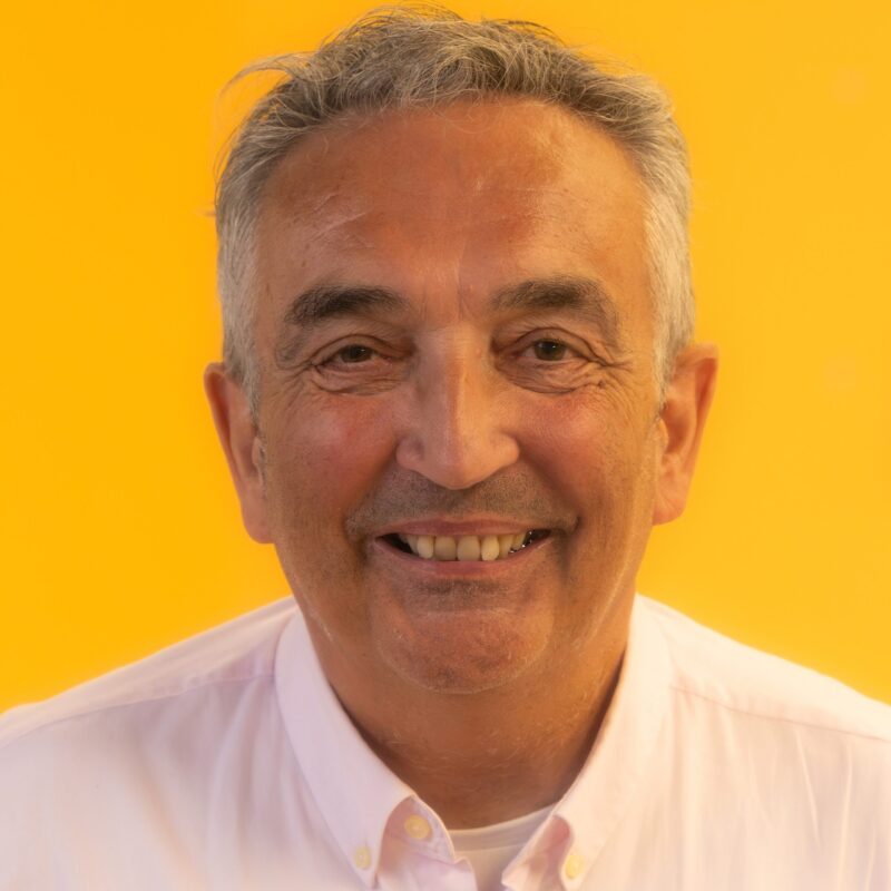 Smiling older man with grey hair wearing a white collared shirt, posed against a bright orange background.