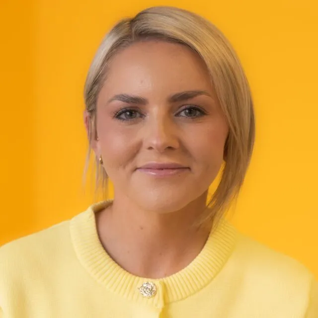 Woman with short blonde hair wearing a yellow jumper, standing in front of a solid yellow background, looking directly at the camera with a soft smile.