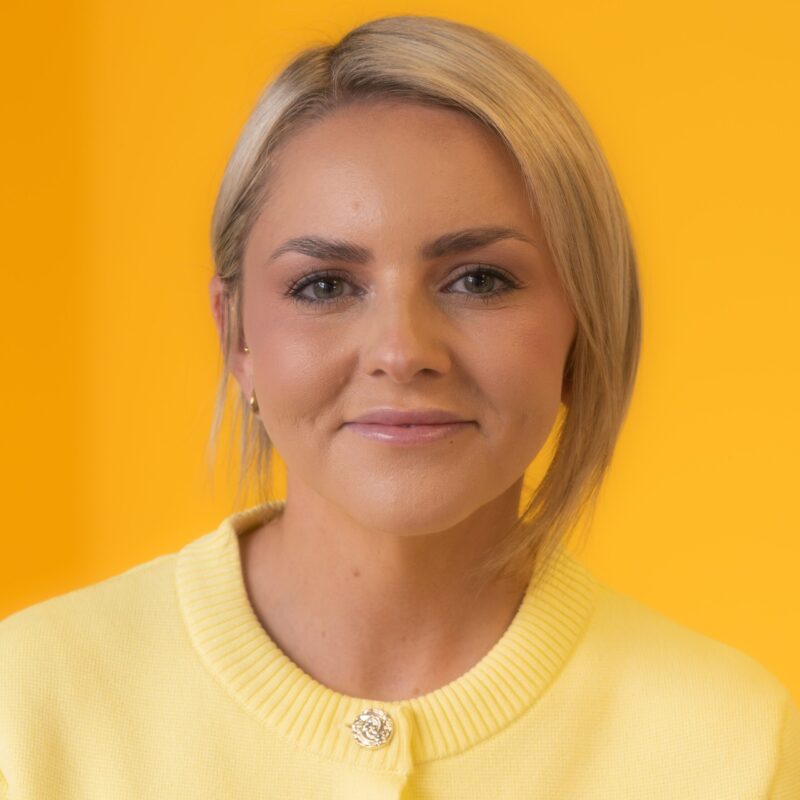 Woman with short blonde hair wearing a yellow jumper, standing in front of a solid yellow background, looking directly at the camera with a soft smile.