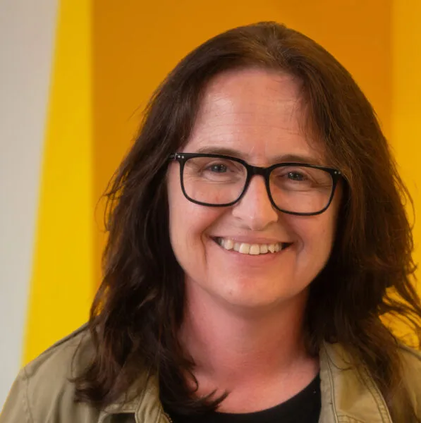 A woman with long brown hair and glasses smiles at the camera, standing in front of a bright yellow background.