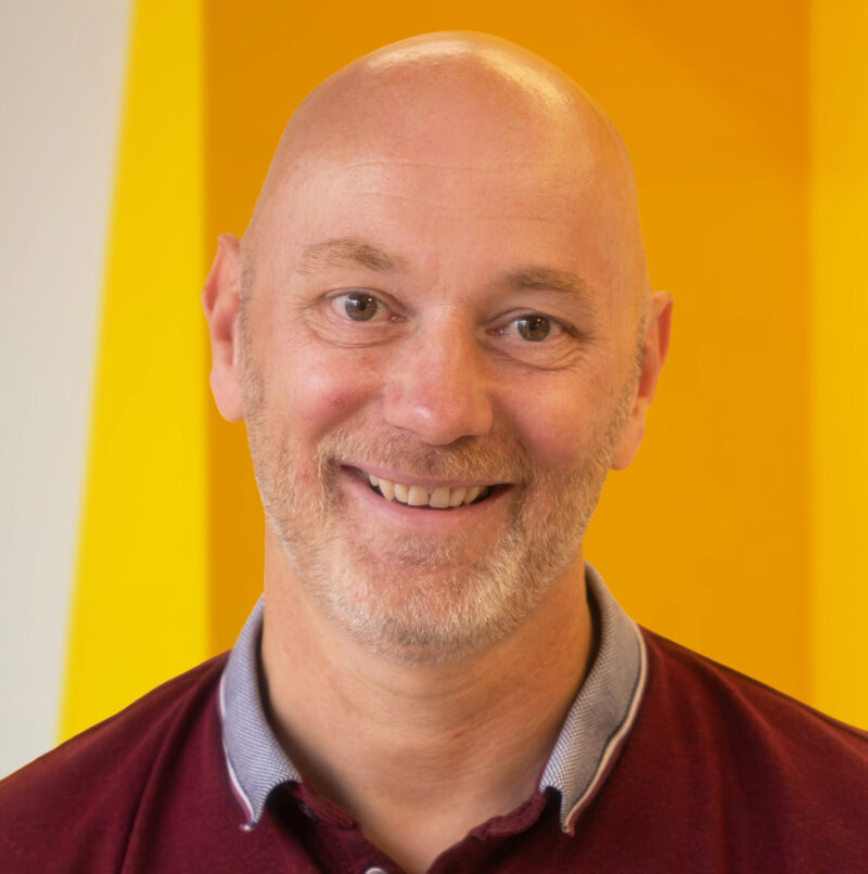 Bald man with a light beard and maroon collared shirt smiles in front of a bright yellow background.