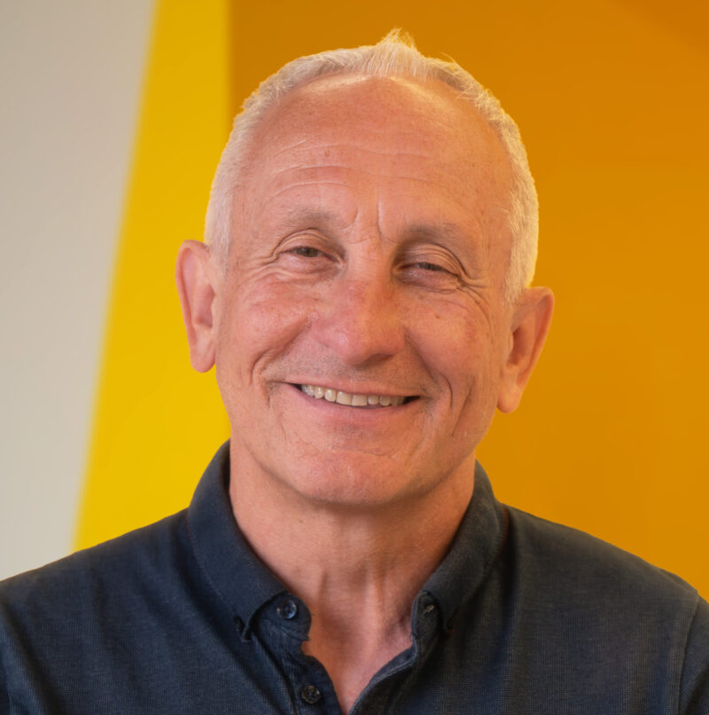 Older man with short grey hair smiling, wearing a dark button-up shirt, standing in front of a yellow and light beige background.