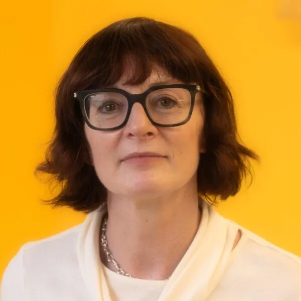 A woman with short brown hair, glasses, and a white top poses in front of a bright yellow background.