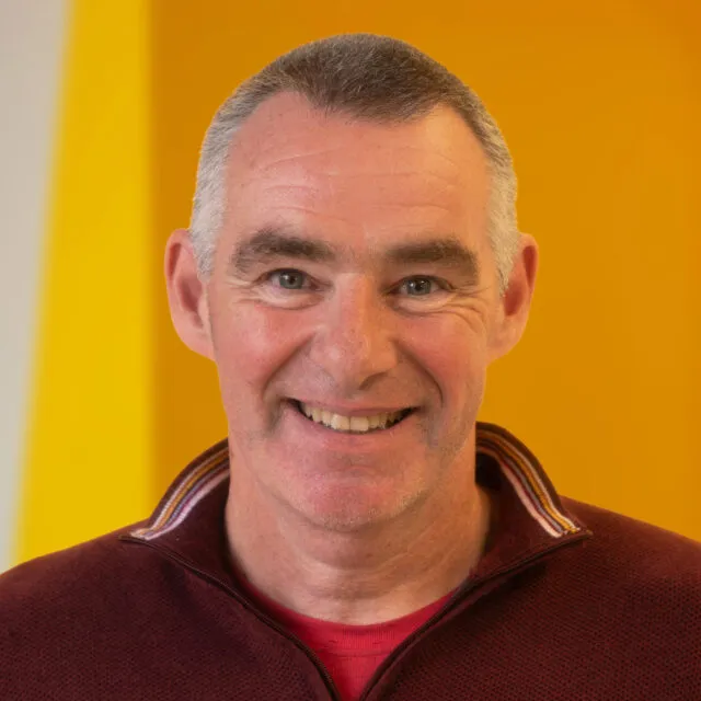 A middle-aged man with short grey hair smiles at the camera, wearing a maroon jumper with a striped collar. The background is bright yellow with a white stripe.