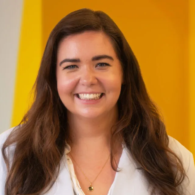 A woman with long brown hair smiles, wearing a white blouse and a gold necklace, standing in front of a bright yellow background.