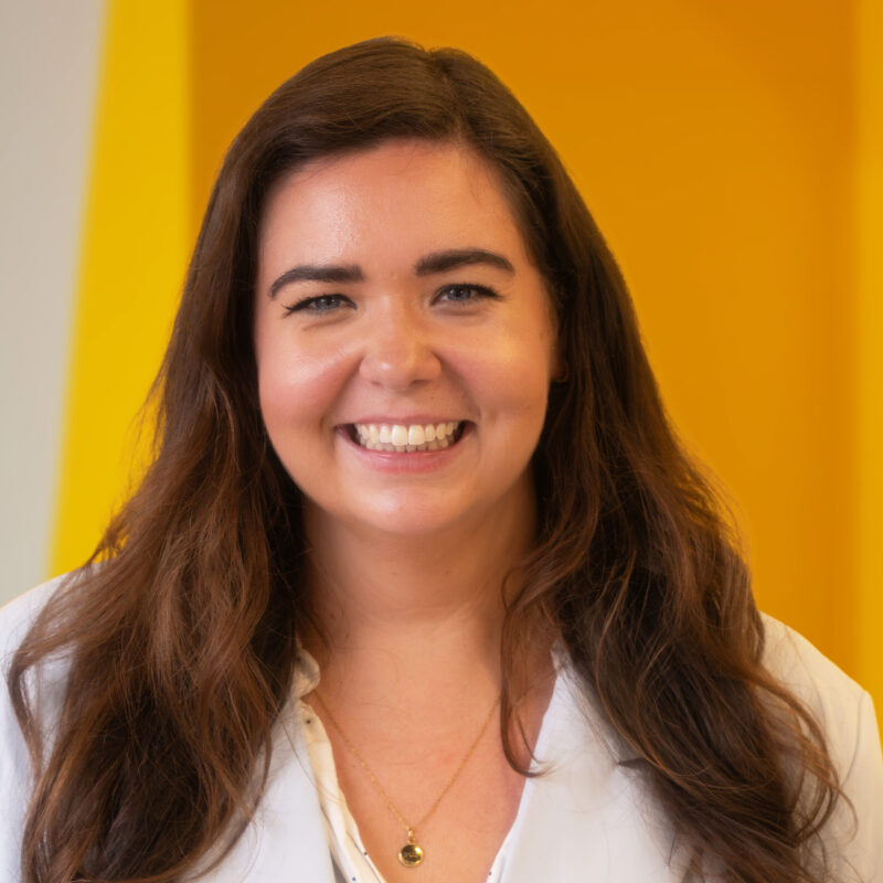 A woman with long brown hair smiles, wearing a white blouse and a gold necklace, standing in front of a bright yellow background.