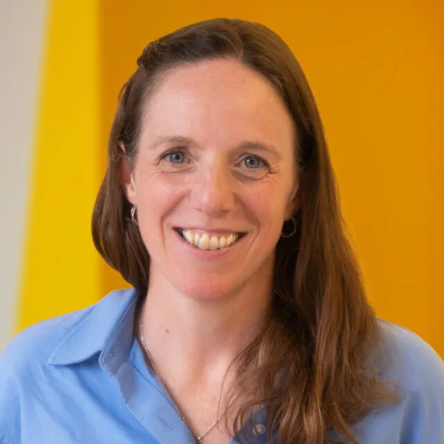 A woman with long brown hair, wearing a light blue shirt and silver hoop earrings, smiles in front of a bright yellow background.