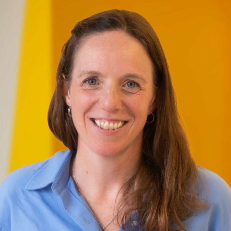 A woman with long brown hair, wearing a light blue shirt and silver hoop earrings, smiles in front of a bright yellow background.