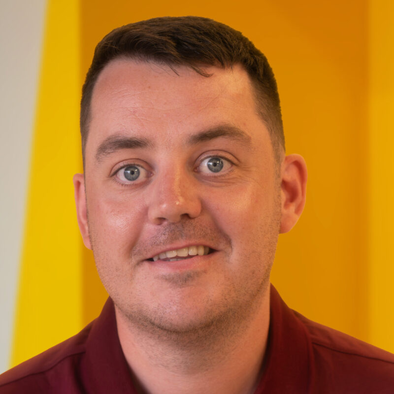 A man with short brown hair and blue eyes smiles slightly, standing in front of a bright yellow background. He wears a maroon collared shirt.