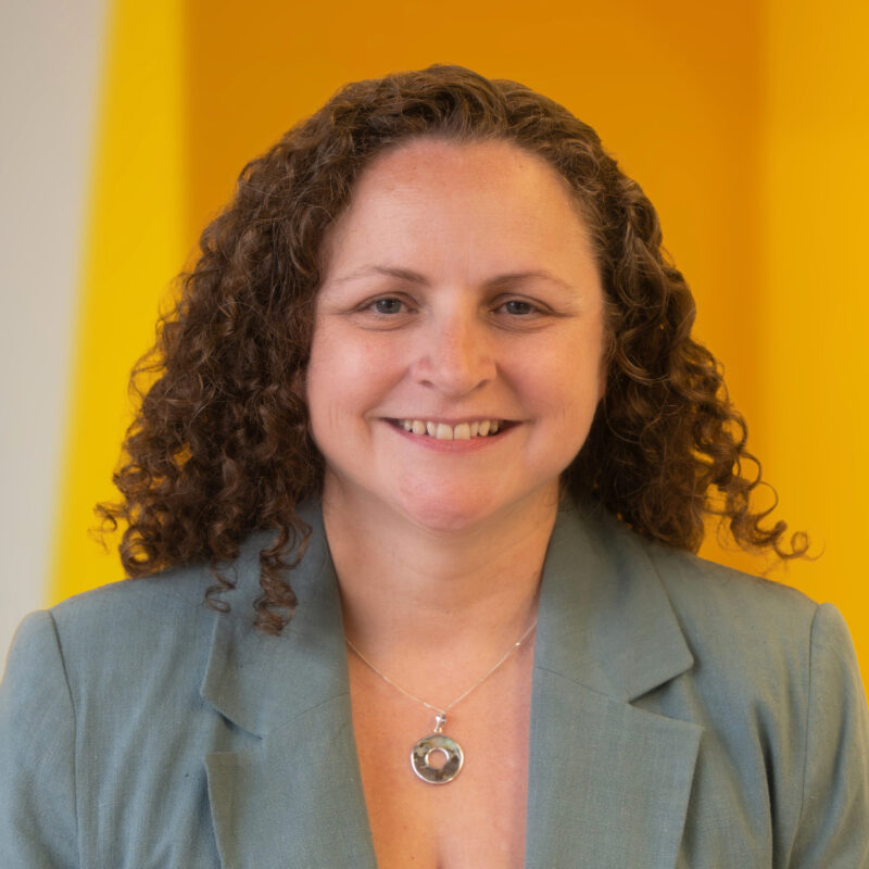 A woman with curly brown hair, wearing a light blue jacket and silver necklace, smiles in front of a bright yellow background.
