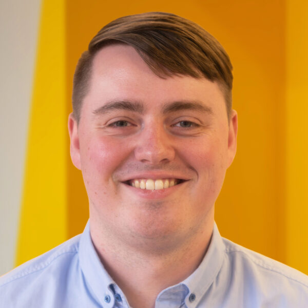 A smiling person with short brown hair wearing a light blue collared shirt stands in front of a bright yellow background.