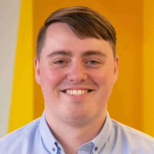 A smiling person with short brown hair wearing a light blue collared shirt stands in front of a bright yellow background.
