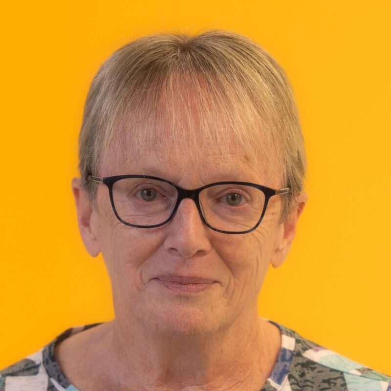 Older adult with short grey hair and black glasses, wearing a patterned shirt, smiling gently in front of a bright yellow background.