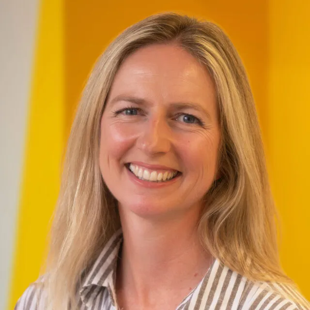 A woman with long blonde hair smiles at the camera, wearing a striped jumper. The background is bright yellow.