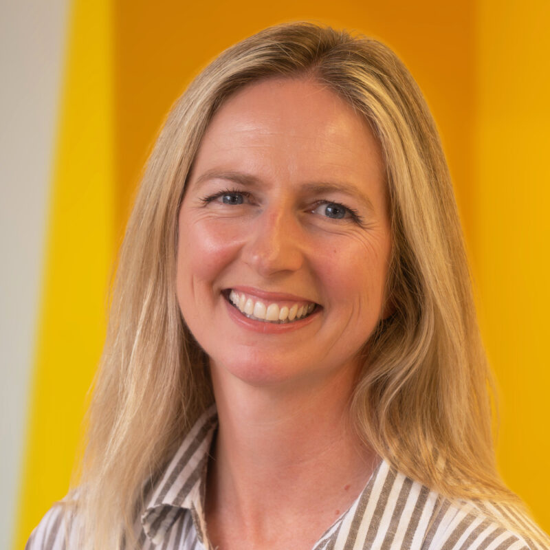 A woman with long blonde hair smiles at the camera, wearing a striped jumper. The background is bright yellow.