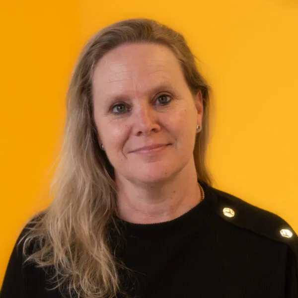 A woman with long blonde hair wearing a black top smiles softly in front of a bright yellow background.