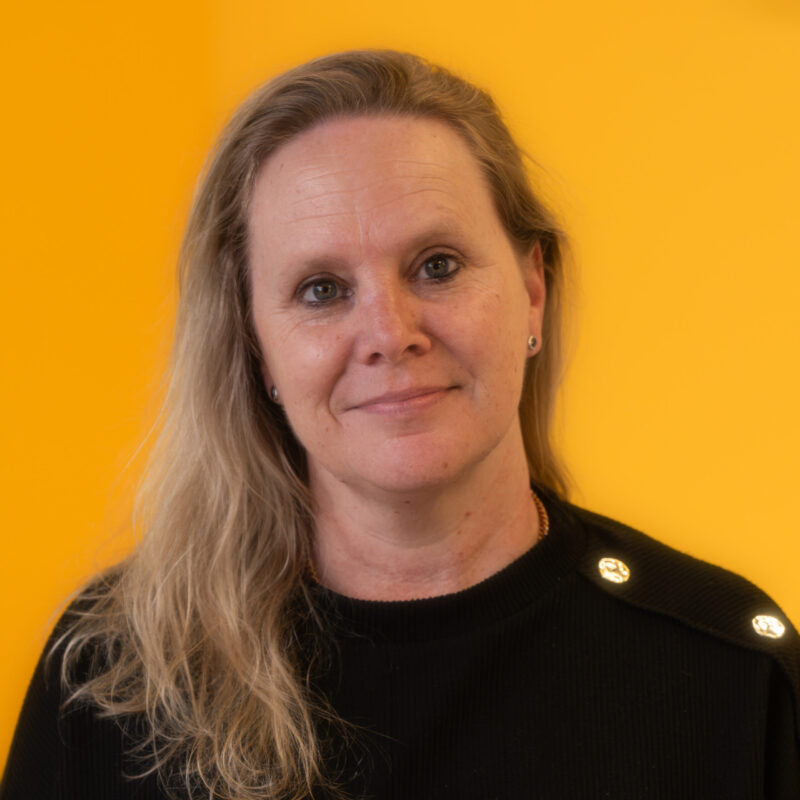 A woman with long blonde hair wearing a black top smiles softly in front of a bright yellow background.
