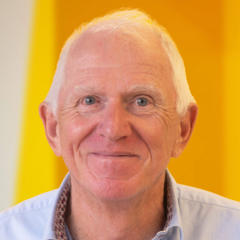 An older man with short white hair and blue eyes is smiling softly, wearing a light blue collared shirt in front of a bright yellow background.