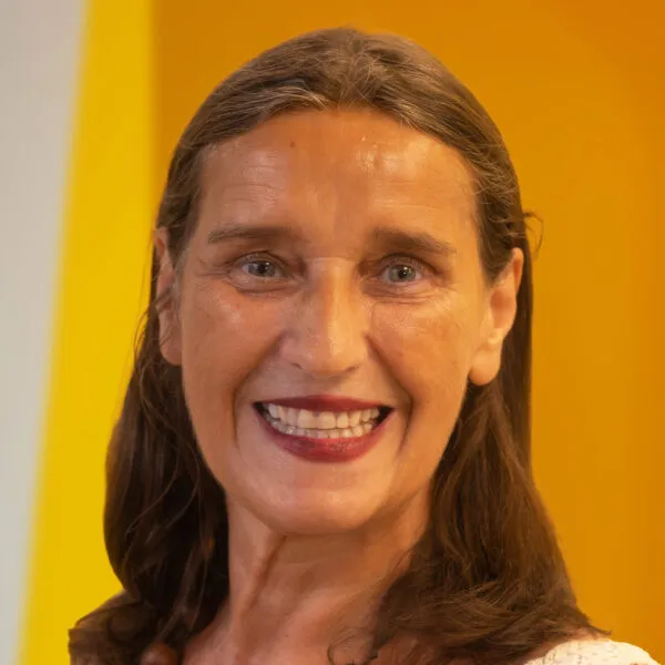 A woman with long brown hair smiles in front of a bright yellow background. She is wearing a light-coloured top and has a cheerful expression.