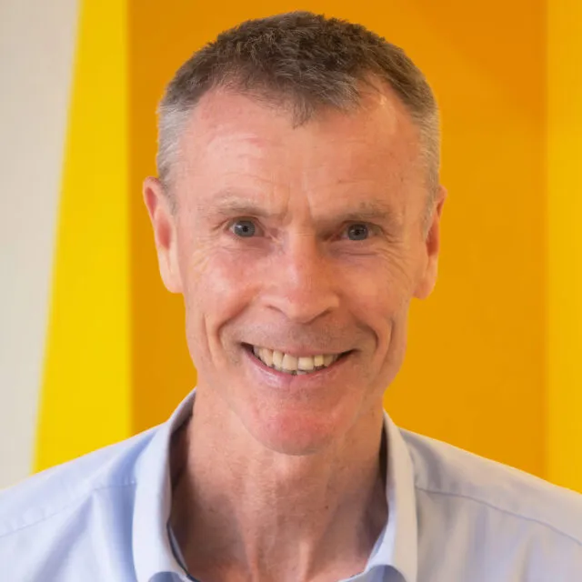 Smiling older man with short grey hair and a light blue collared shirt stands in front of a bright yellow background.