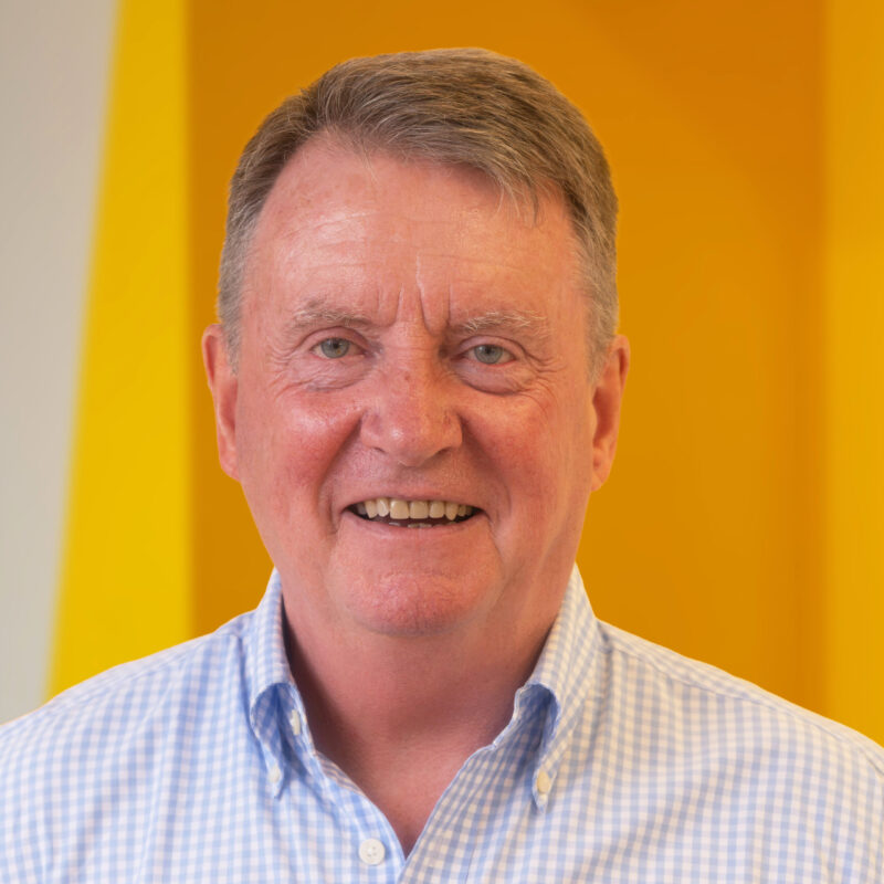 Smiling older man with short grey hair wearing a light blue checked shirt, standing in front of a bright yellow and white background.