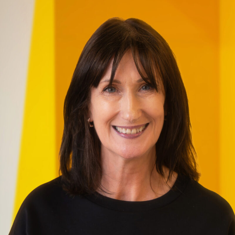 A woman with shoulder-length dark hair and a black top smiles in front of a bright yellow and white background.