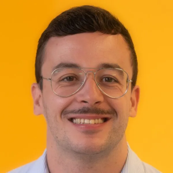 A man with short brown hair, wearing glasses and a light blue shirt, smiles in front of a bright yellow background.