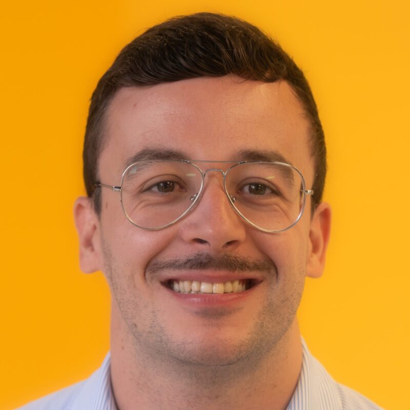 A man with short brown hair, wearing glasses and a light blue shirt, smiles in front of a bright yellow background.