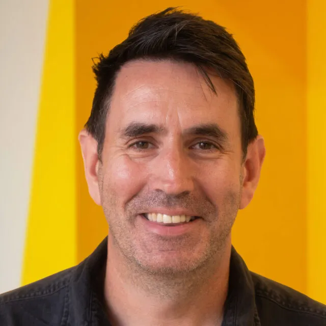 A man with short dark hair smiles at the camera whilst standing in front of a bright yellow background. He is wearing a dark shirt.