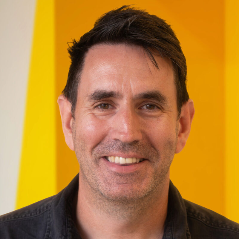 A man with short dark hair smiles at the camera whilst standing in front of a bright yellow background. He is wearing a dark shirt.
