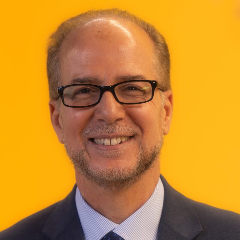 A man wearing glasses and a suit smiles in front of a bright yellow background.