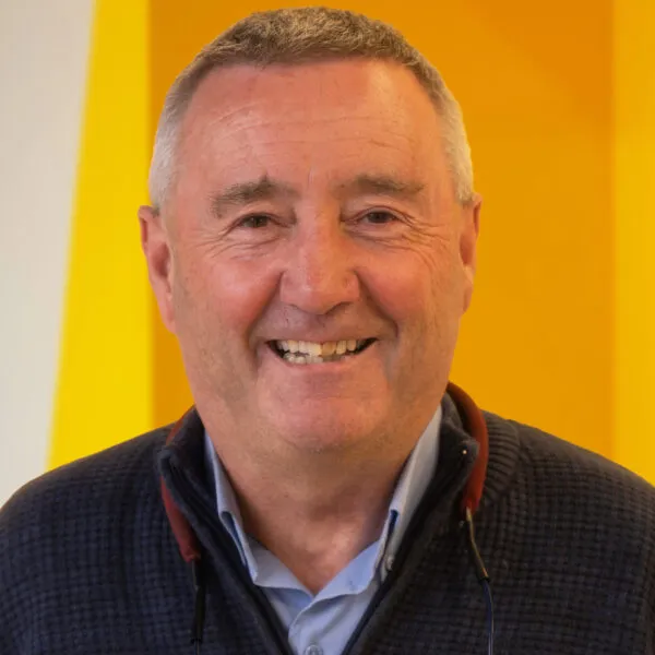 Smiling older man with short grey hair wearing a dark jacket and light blue shirt, standing in front of a bright yellow background.