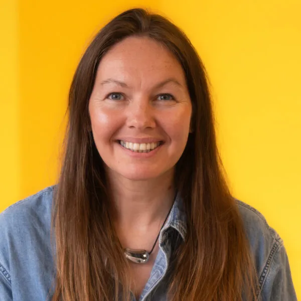 A woman with long brown hair smiles at the camera. She is wearing a denim shirt and a silver necklace, standing in front of a bright yellow background.